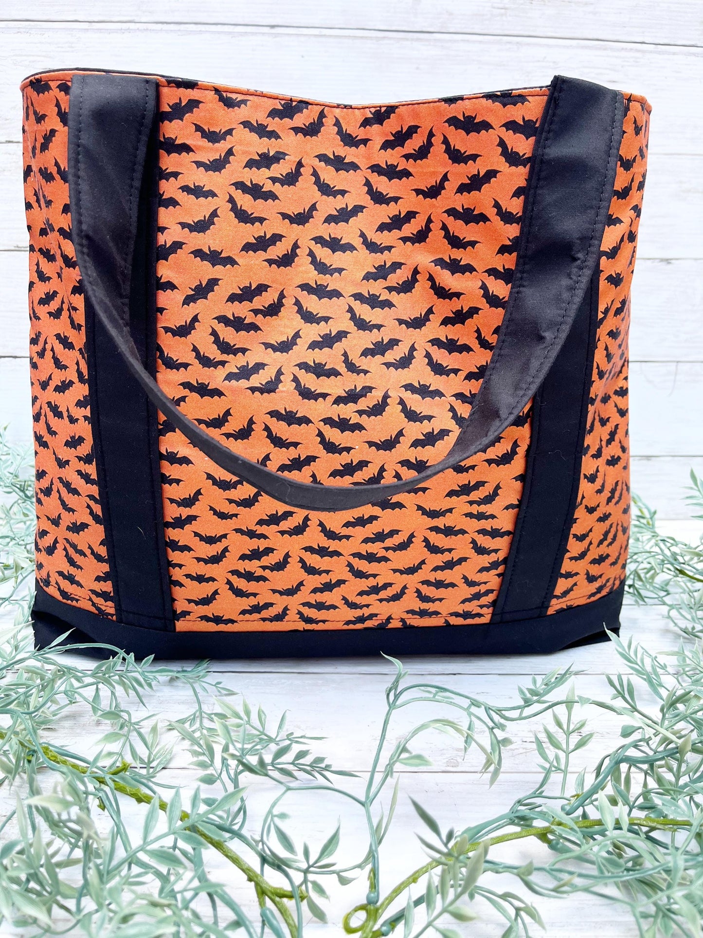 Orange tote bag with black bat pattern on a white wooden surface with greenery.