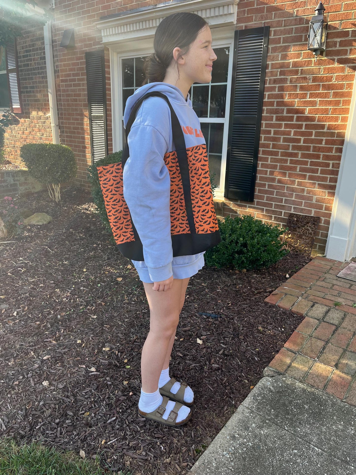 Person wearing  orange and black bat themed totebag standing outside a house.