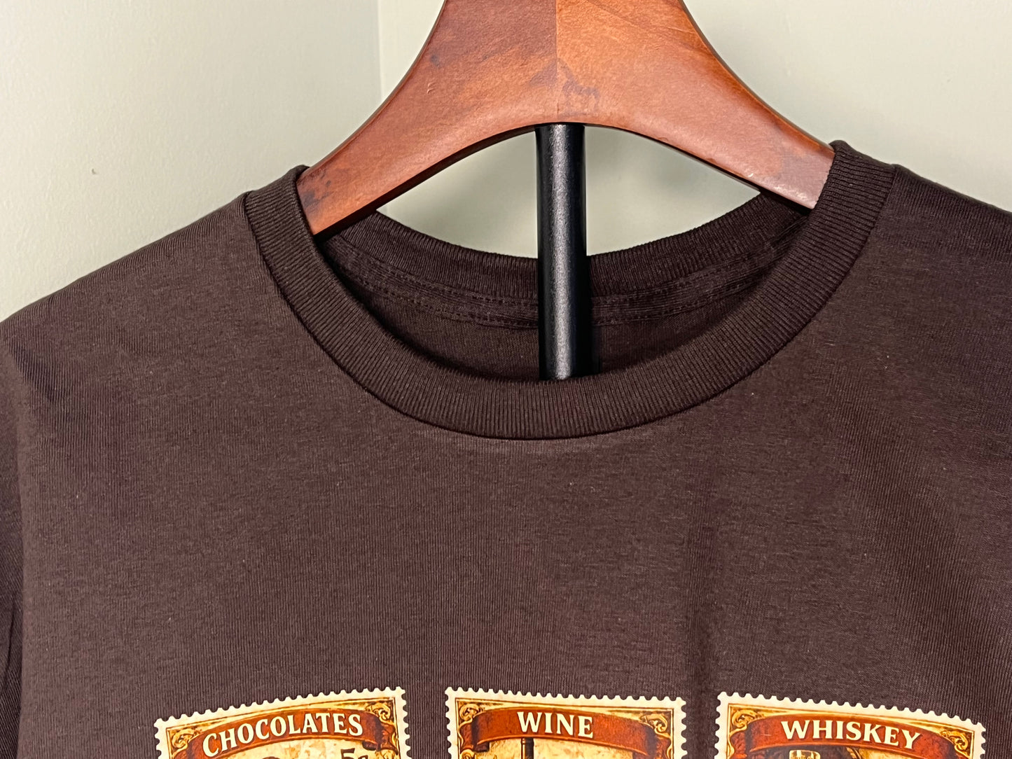 Brown t-shirt with 'Chocolates', 'Wine', and 'Whiskey' vintage stamps  on a wooden hanger.