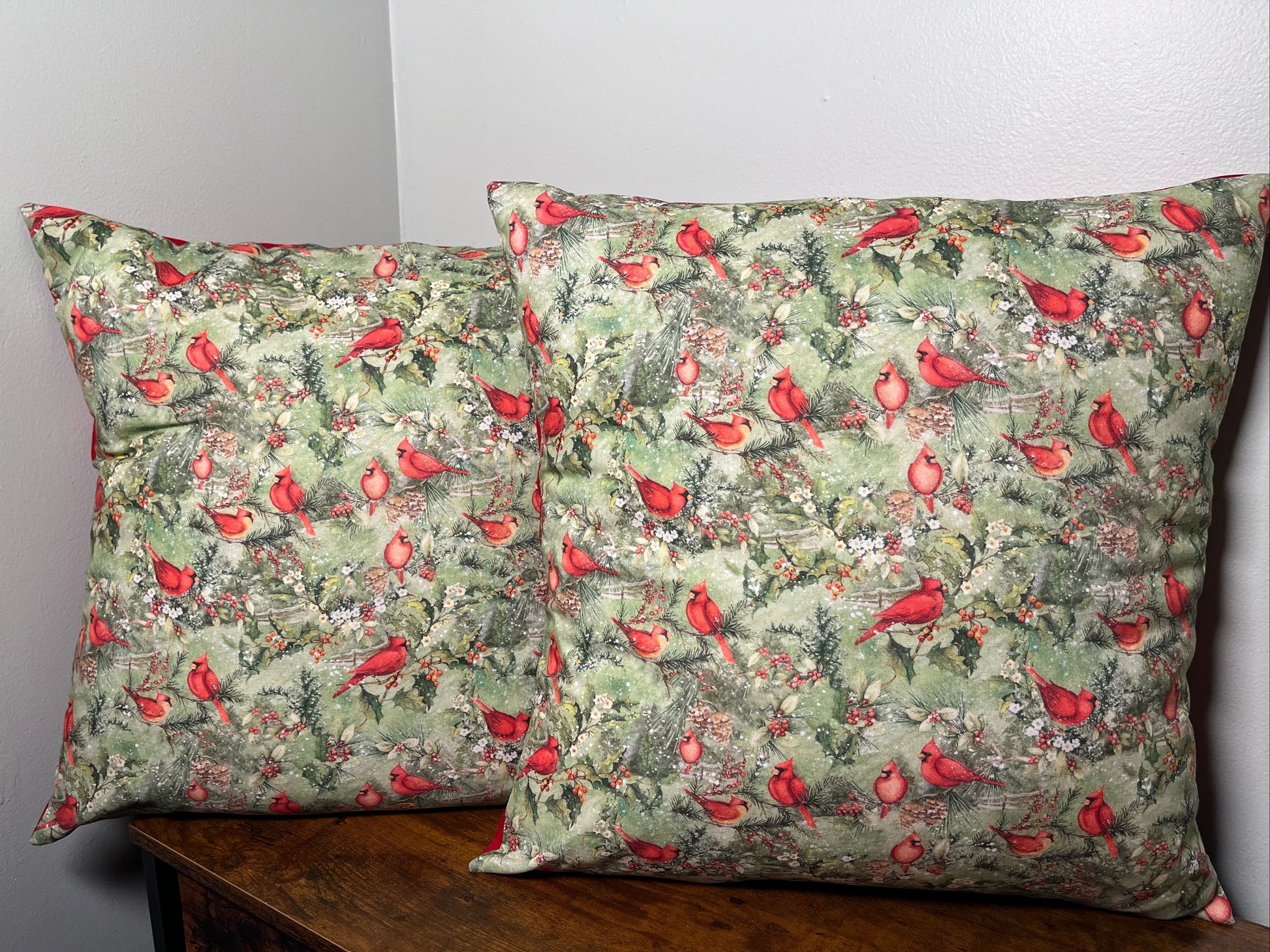 Two Christmas patterned pillows with Cardinals  on a wooden surface against a gray wall.