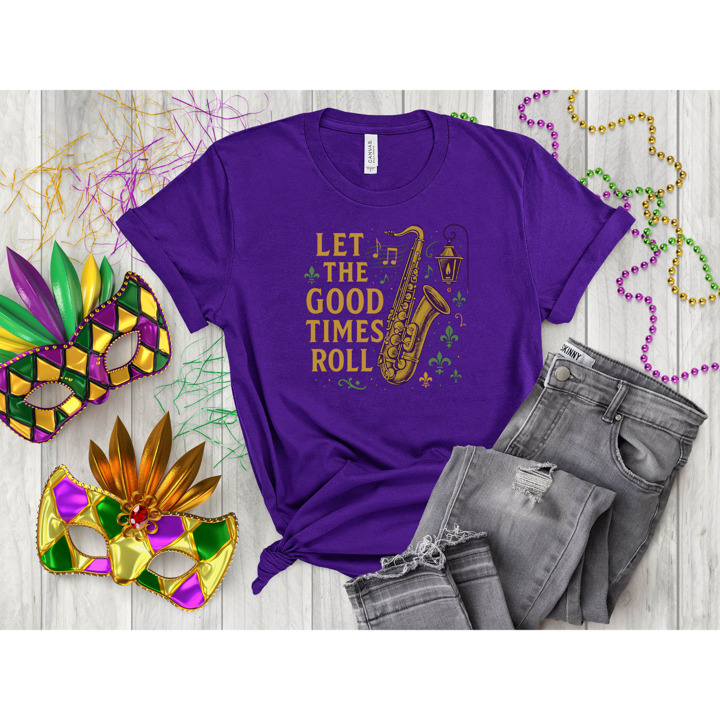 Purple t-shirt with 'Let the Good Times Roll' saxophone design, surrounded by Mardi Gras masks and beads on a wooden surface.